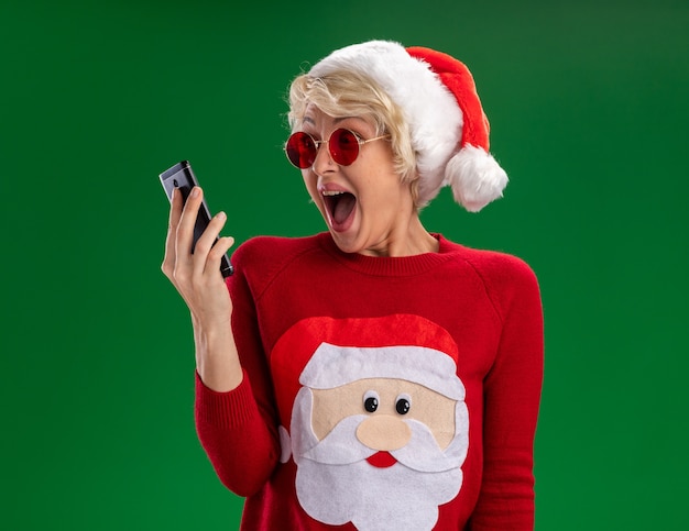 Impresionada joven rubia con sombrero de navidad y suéter de navidad de santa claus con gafas sosteniendo y mirando el teléfono móvil aislado sobre fondo verde