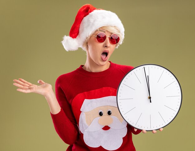 Impresionada joven rubia con sombrero de navidad y suéter de navidad de santa claus con gafas sosteniendo el reloj mirando a la cámara mostrando la mano vacía aislada sobre fondo verde oliva