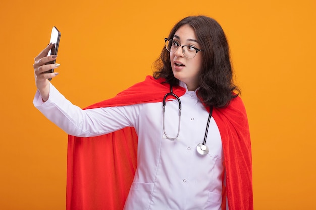 Impresionada joven superhéroe caucásica en capa roja vistiendo uniforme médico y estetoscopio con gafas tomando selfie aislado en pared naranja