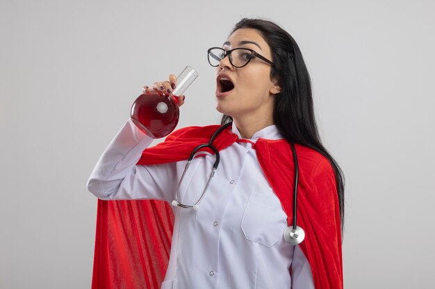 Impresionada joven superhéroe caucásica con gafas y estetoscopio sosteniendo frasco químico con líquido rojo tratando de beberlo mirando a cámara aislada sobre fondo blanco