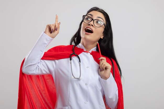 Impresionada joven superhéroe caucásica con gafas sosteniendo un lápiz rojo mirando al lado apuntando hacia arriba aislado en la pared blanca con espacio de copia