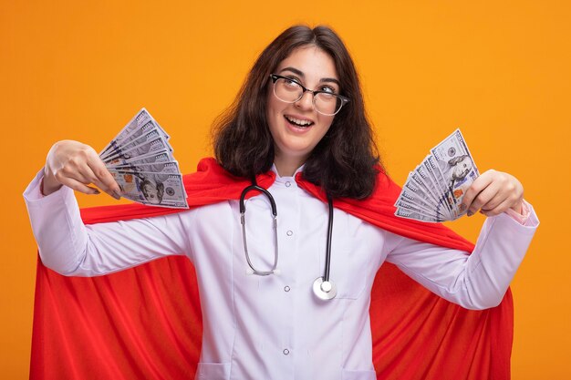 Impresionada joven superhéroe caucásica vestida con uniforme médico y un estetoscopio con gafas sosteniendo dinero mirando hacia arriba aislado en la pared