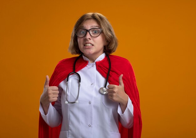 Impresionada joven superhéroe rubia en capa roja con uniforme de médico y gafas con estetoscopio mirando al frente mostrando los pulgares para arriba aislados en la pared con espacio de copia