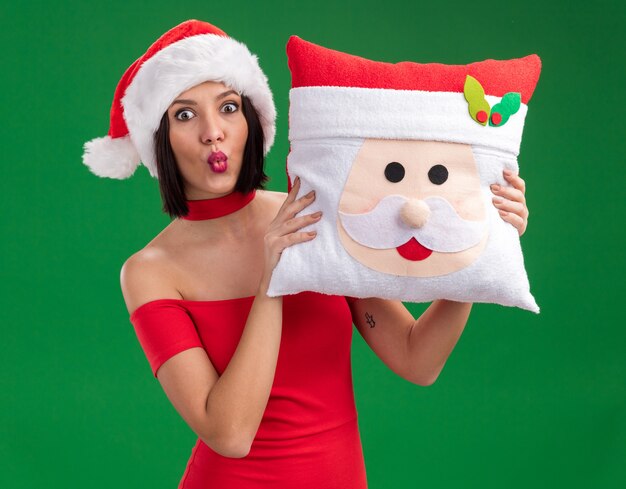 Impresionada joven vistiendo gorro de Papá Noel con almohada de santa claus con labios fruncidos aislados en la pared verde