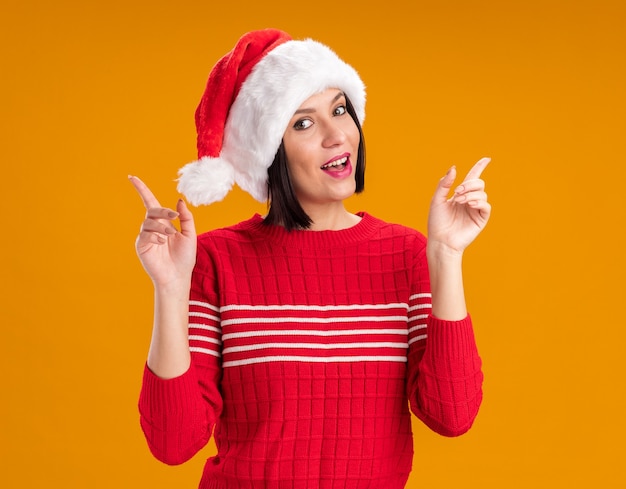 Impresionada joven vistiendo gorro de Papá Noel apuntando hacia arriba aislado en la pared naranja