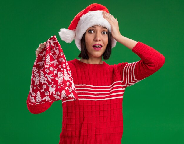 Impresionada joven vistiendo gorro de Papá Noel con saco de regalo de navidad manteniendo la mano en la cabeza aislada en la pared verde