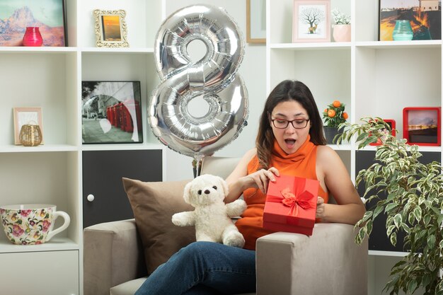 Impresionada jovencita con gafas ópticas abriendo y mirando la caja de regalo sentada en un sillón en la sala de estar en el día internacional de la mujer de marzo