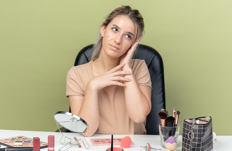 Foto gratuito impresionada mirando lado hermosa joven sentada en el escritorio con herramientas de maquillaje poniendo la mano en la mejilla aislada en la pared verde oliva