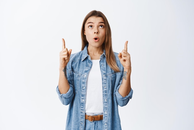 Impresionada modelo bastante femenina apuntando con el dedo hacia arriba y levantando la cabeza en el anuncio superior mirando algo fascinado de pie contra el fondo blanco