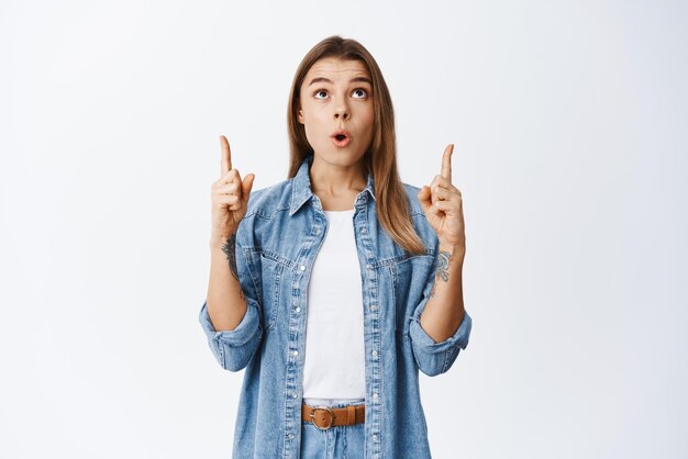 Impresionada modelo bastante femenina apuntando con el dedo hacia arriba y levantando la cabeza en el anuncio superior mirando algo fascinado de pie contra el fondo blanco