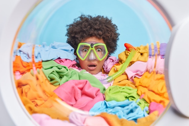 Foto gratuita impresionada mujer afroamericana de pelo rizado mira fijamente maravillada a través de gafas de esnórquel enterrada en ropa multicolor lava la ropa con regularidad