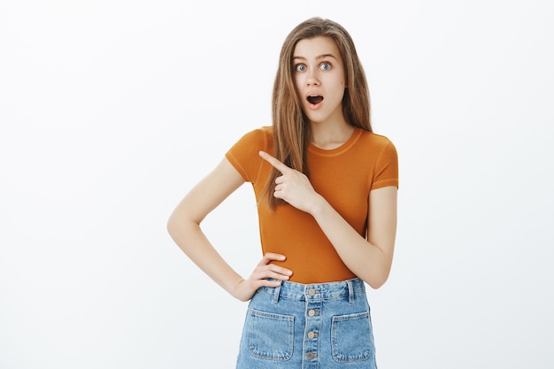 Impresionada mujer atractiva que señala con el dedo a la izquierda, invitando a cursos en línea o visitar la tienda