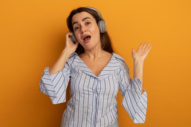 Impresionada mujer bonita en auriculares se encuentra con la mano levantada aislada en la pared naranja