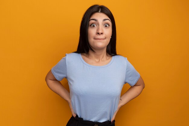 Impresionada mujer bonita morena pone las manos en la cintura y mira al frente aislado en la pared naranja