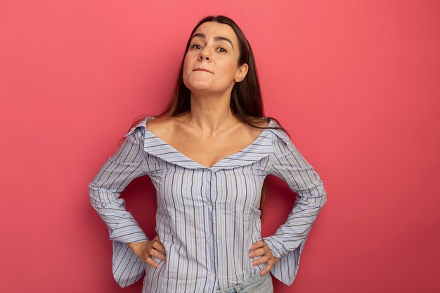 Impresionada mujer bonita pone las manos en la cintura y mira al frente aislado en la pared rosa