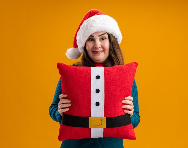 Impresionada mujer caucásica adulta con gorro de Papá Noel y corbata de santa sosteniendo una almohada decorada aislada en la pared naranja con espacio de copia