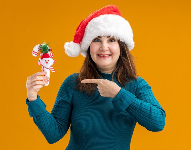 Impresionada mujer caucásica adulta con gorro de Papá Noel sostiene y apunta al bastón de caramelo aislado sobre fondo naranja con espacio de copia