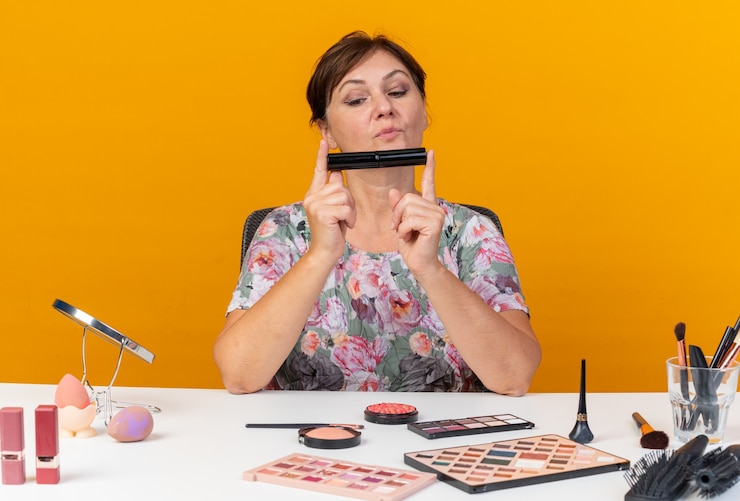Foto gratuito impresionada mujer caucásica adulta sentada a la mesa con herramientas de maquillaje sosteniendo y mirando rímel aislado en la pared naranja con espacio de copia
