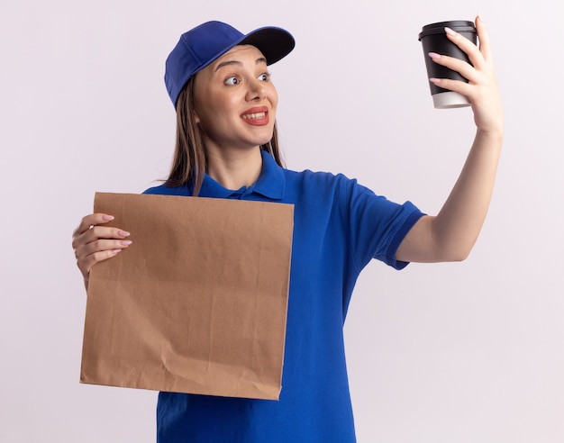 Impresionada mujer de entrega bonita en uniforme sostiene el paquete de papel y mira la taza de papel aislada en la pared blanca con espacio de copia