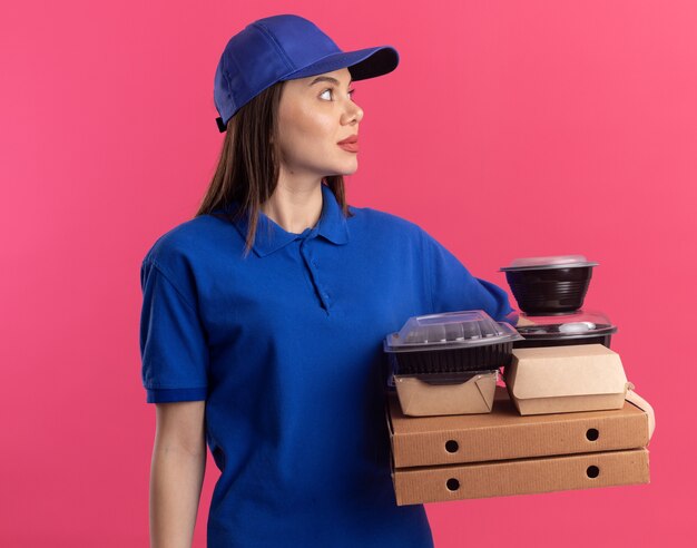 Impresionada mujer de entrega bonita en uniforme sostiene paquetes de alimentos y contenedores en cajas de pizza mirando al lado en rosa