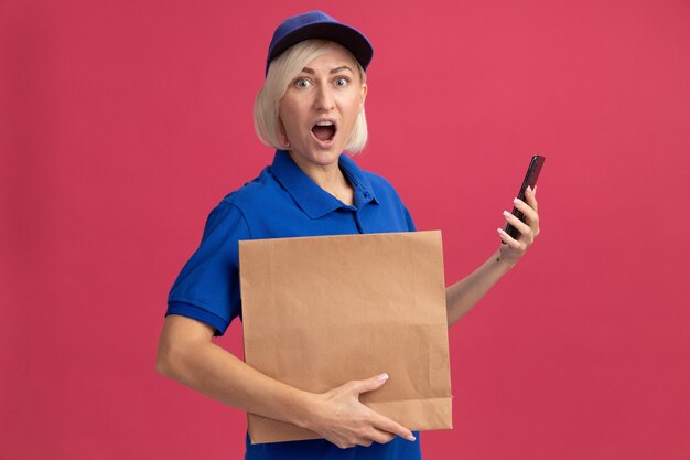 Impresionada mujer de entrega rubia de mediana edad en uniforme azul y gorra con paquete de papel y teléfono móvil aislado en la pared rosa