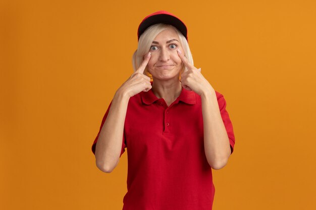 Impresionada mujer de entrega rubia de mediana edad en uniforme rojo y gorra tirando hacia abajo los párpados frunciendo los labios