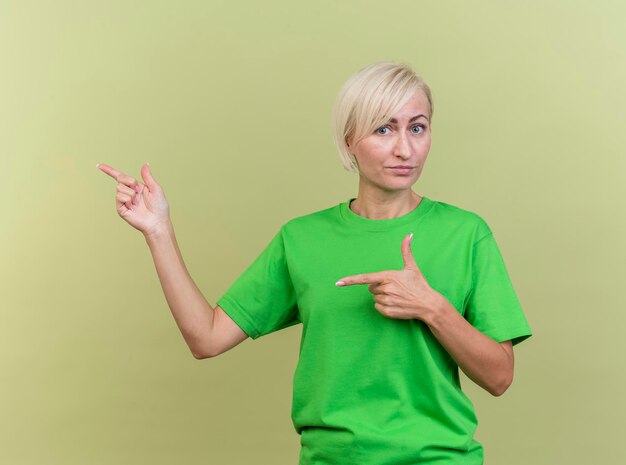 Impresionada mujer eslava rubia de mediana edad mirando al frente apuntando al lado aislado en la pared verde oliva