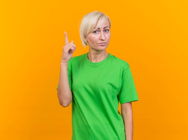 Impresionada mujer eslava rubia de mediana edad mirando al frente apuntando hacia arriba aislado en la pared amarilla con espacio de copia