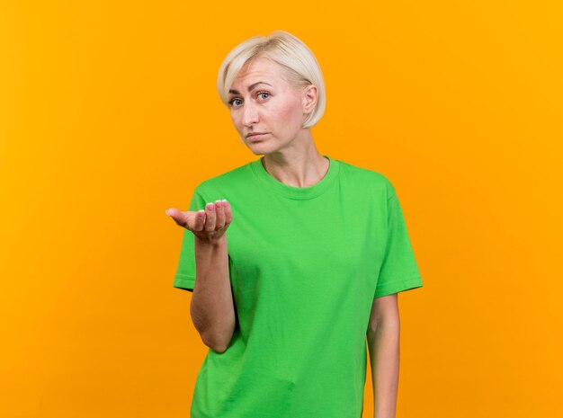 Impresionada mujer eslava rubia de mediana edad mirando al frente mostrando la mano vacía aislada en la pared amarilla con espacio de copia