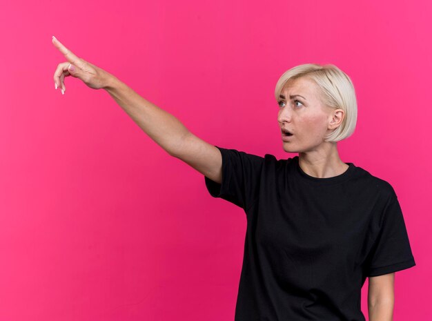 Impresionada mujer eslava rubia de mediana edad mirando y apuntando al lado aislado en la pared rosa