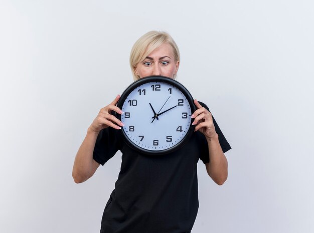 Impresionada mujer eslava rubia de mediana edad sosteniendo y mirando el reloj aislado sobre fondo blanco con espacio de copia