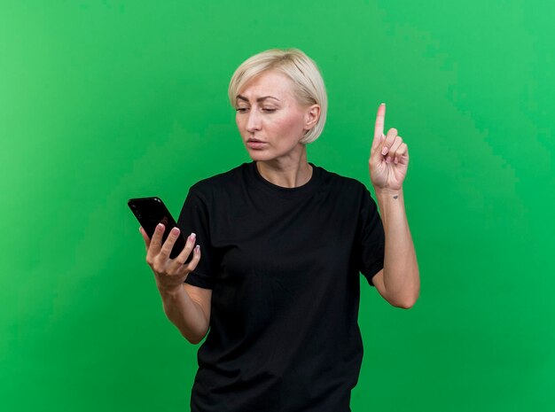 Impresionada mujer eslava rubia de mediana edad sosteniendo y mirando el teléfono móvil levantando el dedo aislado en la pared verde con espacio de copia