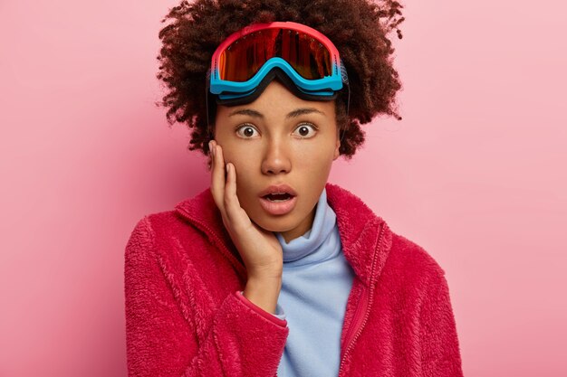 Impresionada mujer de invierno lleva gafas para hacer snowboard, jersey rojo y cuello alto azul, mira con sorpresa, posa sobre fondo rosa