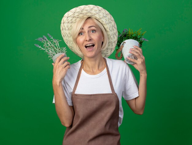 Impresionada mujer jardinero rubia de mediana edad en uniforme con sombrero sosteniendo macetas cerca de la cabeza mirando al lado aislado en la pared verde