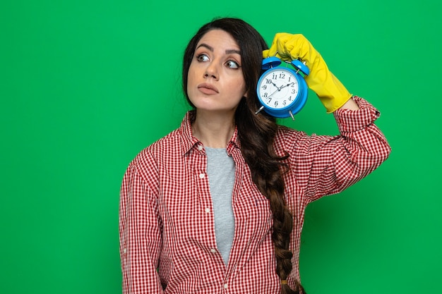 Impresionada mujer limpiadora bastante caucásica con guantes de goma sosteniendo el despertador y mirando al lado