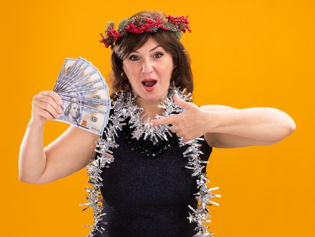 Impresionada mujer de mediana edad con corona de Navidad y guirnalda de oropel alrededor del cuello sosteniendo y apuntando al dinero mirando a cámara aislada sobre fondo naranja