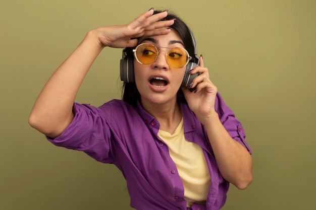 Foto gratuita impresionada mujer morena bonita en gafas de sol con auriculares mantiene la palma en la frente mirando al lado aislado en la pared verde oliva