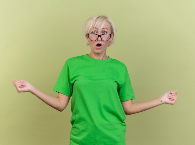 Impresionada mujer rubia de mediana edad con gafas mirando al frente estirando los puños aislados en la pared verde oliva