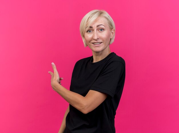 Impresionada mujer rubia de mediana edad mirando al frente apuntando detrás aislado en la pared rosa