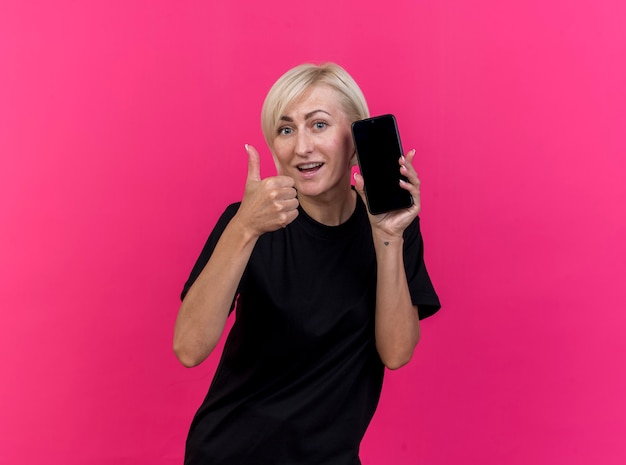 Impresionada mujer rubia de mediana edad que muestra el teléfono móvil y el pulgar hacia arriba mirando al frente aislado en la pared rosa