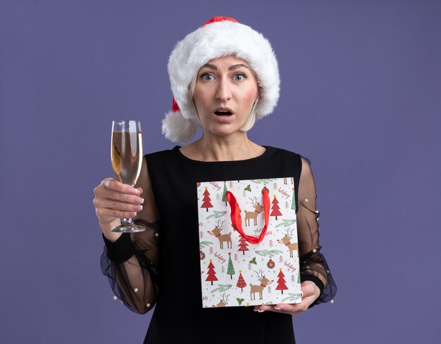 Impresionada mujer rubia de mediana edad con sombrero de navidad mirando a la cámara sosteniendo una copa de champán y una bolsa de regalo de navidad aislada sobre fondo púrpura