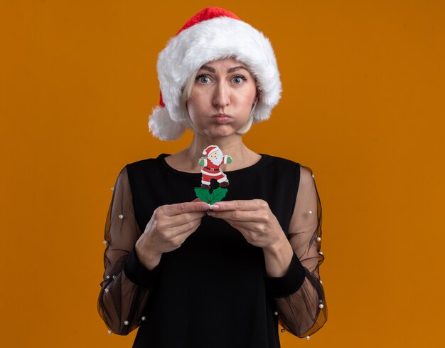 Impresionada mujer rubia de mediana edad con sombrero de navidad sosteniendo el juguete de santa claus hinchando las mejillas aisladas en la pared naranja con espacio de copia