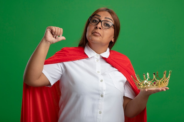 Impresionada mujer superhéroe de mediana edad con gafas sosteniendo puntos de corona a sí misma aislada sobre fondo verde