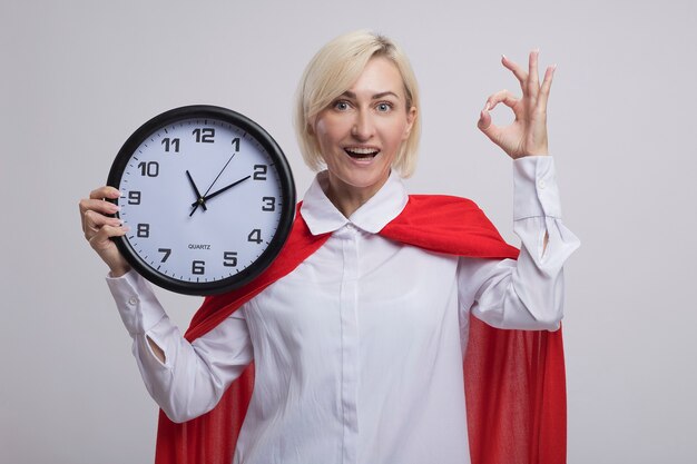 Impresionada mujer de superhéroe rubia de mediana edad en capa roja sosteniendo el reloj mirando al frente haciendo bien signo aislado en la pared blanca