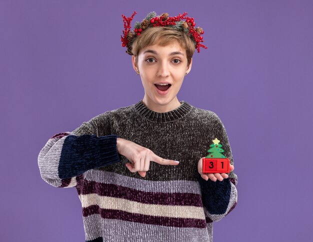 Impresionada niña bonita con corona de cabeza de navidad sosteniendo el juguete del árbol de navidad con fecha apuntando a él aislado en la pared púrpura