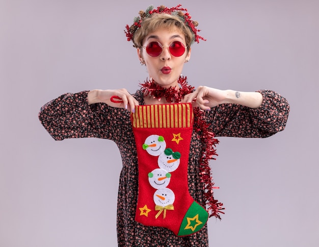 Foto gratuita impresionada niña bonita con corona de navidad y guirnalda de oropel alrededor del cuello con gafas sosteniendo calcetines de navidad mirando al lado aislado sobre fondo blanco.