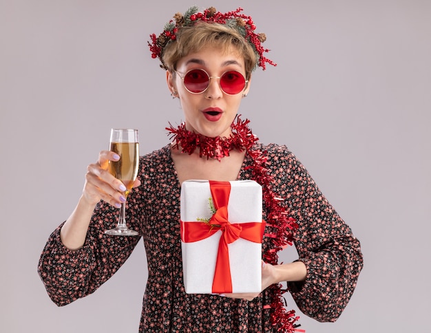 Impresionada niña bonita con corona de Navidad y guirnalda de oropel alrededor del cuello con gafas sosteniendo paquete de regalo y copa de champán mirando a cámara aislada sobre fondo blanco