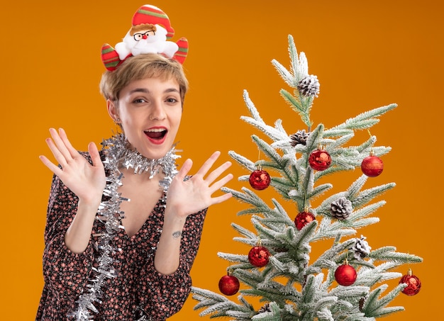 Foto gratuita impresionada niña bonita con diadema de santa claus y guirnalda de oropel alrededor del cuello de pie cerca del árbol de navidad decorado mostrando las manos vacías aisladas en la pared naranja