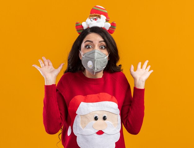 Impresionada niña bonita con diadema de santa claus y un suéter con máscara protectora que muestra las manos vacías aisladas en la pared naranja