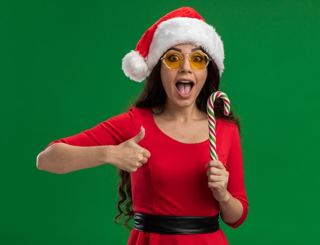 Impresionada niña bonita con gorro de Papá Noel y gafas con bastón de caramelo de Navidad mirando a la cámara mostrando el pulgar hacia arriba aislado sobre fondo verde